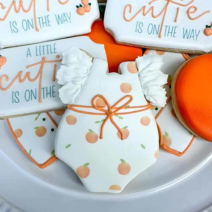 Decorated sugar cookies for baby shower, featuring onesie, cutie plaques, and orange cookies.