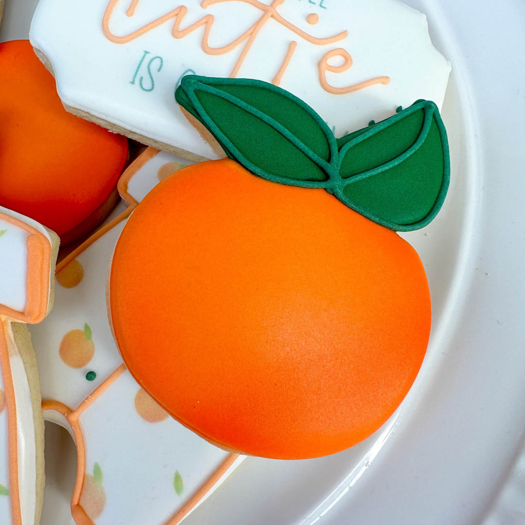 Colorful orange sugar cookie with green leaf, perfect for a baby shower themed 'Little Cutie' collection.