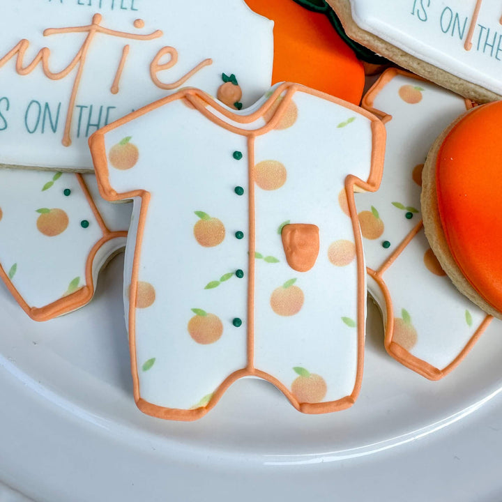 Adorable baby shower cookie decorated as a onesie with orange fruit design, perfect for a Little Cutie theme.