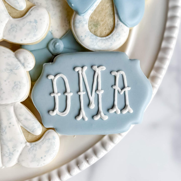 Baby cookie decorated with 'DMA' in white icing on a blue plaque, part of an adorable boy's cookie collection.