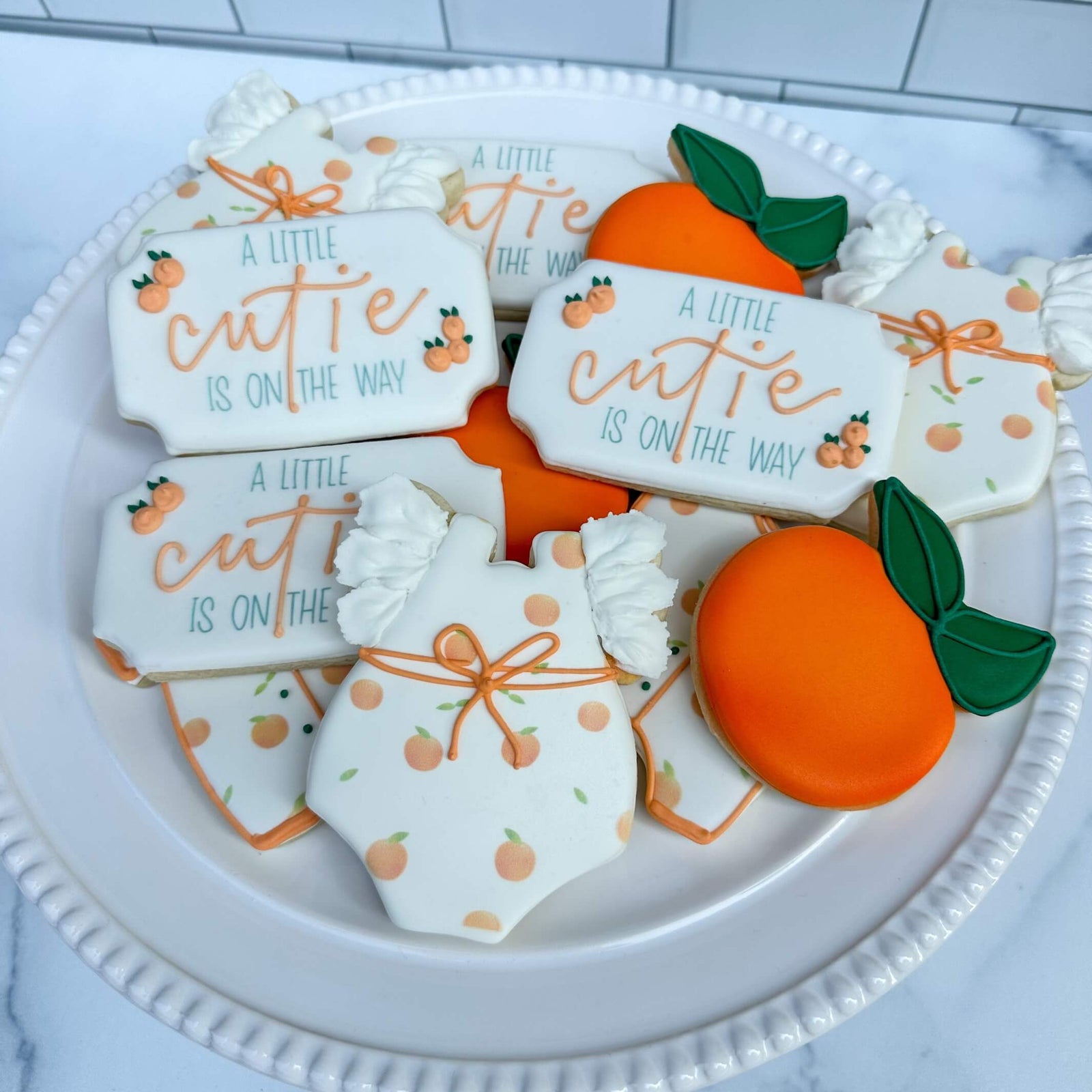 Adorable baby shower cookie set featuring onesies and orange cookies, perfect for a 'Little Cutie' celebration.